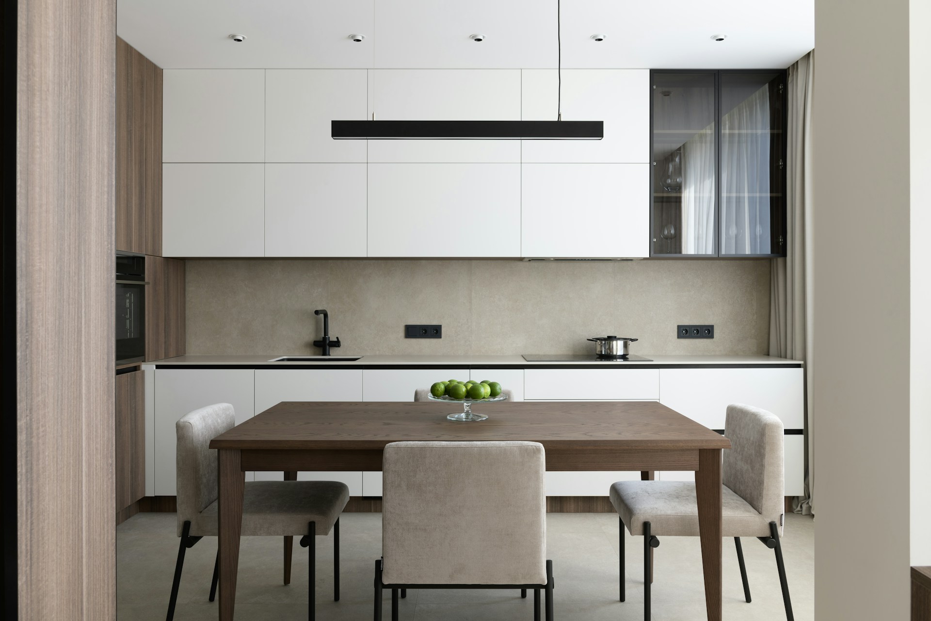 Modern kitchen with white cabinets, wooden dining table, four gray upholstered chairs, bowl of green apples, black sink faucet, stovetop with pot, and a rectangular pendant light.
