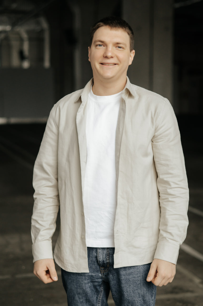 Smiling man wearing a light beige shirt over a white t-shirt and blue jeans standing indoors.