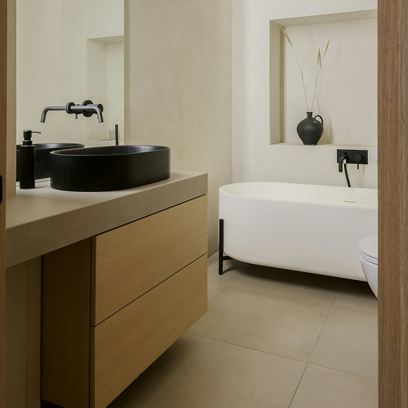 Minimalist bathroom with a black countertop basin, wall-mounted faucet, wooden vanity, and a freestanding white bathtub with a black faucet and a small vase with dried plants in a recessed shelf.